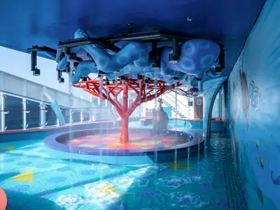 Der farbenfrohe Aurora Borealis Aquapark an Bord der MSC World Europa. Blaue Decke, rote Baumstruktur als Wasserspiel, Pools und Unterwassermalereien an der Wand laden zum Spielen ein.