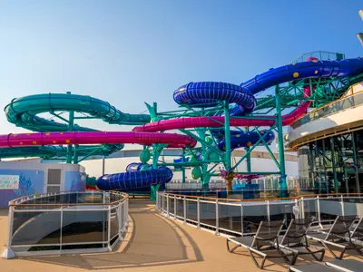 Blick auf den Aurora Borealis Aquapark an Bord der MSC World Europa bei Tageslicht. Verschiedene bunte Wasserrutschen in Blau, Türkis und Pink schlängeln sich über das Deck. Unten rechts sind leere Liegestühle auf dem Sonnendeck zu sehen.