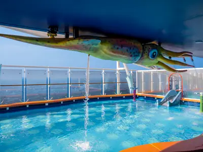 Ein bunter Kraken-Wasserspielbereich mit Pool an Bord der MSC World Europa unter blauem Himmel mit Meerblick. Wasser spritzt aus dem Kraken und kleinen Fontänen. Ein kleiner Kinderrutsche ist zu sehen.