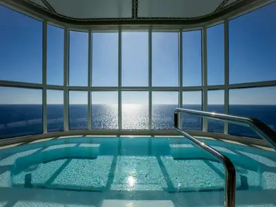 Blick auf einen glitzernden Innenpool an Bord der MSC World Europa mit Panoramablick auf das weite, blaue Meer durch eine Glasfront bei strahlendem Sonnenschein. Ein Geländer fuehrt ins tuerkisfarbene Wasser.