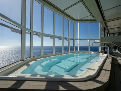 Ein heller Innenpool an Bord der MSC World Europa mit weitem Blick auf das sonnenbeschienene Meer durch grosse Panoramafenster. Das Poolwasser ist türkisblau und die Umrandung weiss. Ohne Personen.