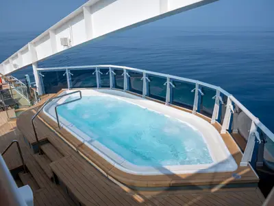 Ein blauer, sprudelnder Whirlpool auf dem Holzdeck der MSC World Europa. Im Hintergrund erstreckt sich das weite, blaue Meer unter klarem Himmel. Ein gläsernes Geländer trennt den Deckbereich vom Ozean.