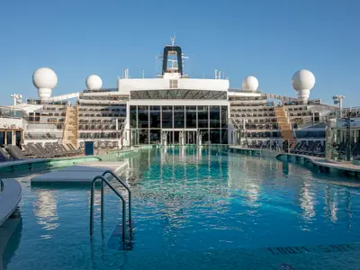 Blick auf den weitläufigen La Plage Poolbereich der MSC World Europa. Blaue Wasserflächen, umgeben von Liegestühlen und der modernen Glasarchitektur des Schiffsinneren unter klarem Himmel.