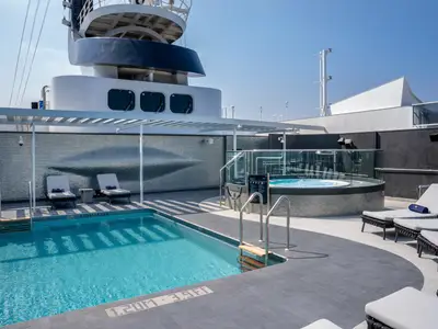 Ein weitläufiges Pooldeck an Bord der MSC World Europa mit einem rechteckigen Pool, einem runden Whirlpool und zahlreichen Sonnenliegen unter blauem Himmel. Eine moderne Pergola spendet Schatten.