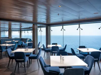 Ein modernes Restaurant an Bord der MSC World Europa mit blauen Samtsesseln und weißen Tischen, umgeben von großen Glasfenstern, die einen weiten Blick auf das blaue Meer bieten. Die Holzdecke spiegelt sich leicht im Glas.