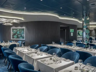Ein elegantes, modernes Restaurant, genannt 'Bubbles', an Bord der MSC World Europa. Es sind zahlreiche Tische mit blauen Samtstuehlen zu sehen, die festlich mit weissen Tischdecken und Geschirr gedeckt sind. Die Waende sind mit dunklem Holz verkleidet und zeigen grosse Kunstwerke mit blauen Blasenmotiven. Eine spiegelnde Saeule mit blauen Akzenten praegt das Ambiente, waehrend die Decke mit indirekter Beleuchtung und stilvollen Lampen versehen ist.