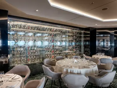Elegantes Restaurant an Bord der MSC World Europa. Gedeckte Tische vor einer grossen, beleuchteten Glasweinwand. Moderne Einrichtung mit hellen Stühlen. Ohne Personen.