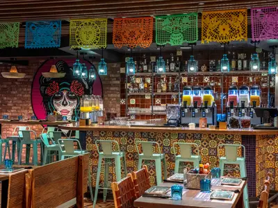 Vibrantes Spezialitätenrestaurant HOLA! Tacos & Cantina an Bord der MSC World Europa. Eine Bar mit Getränkespendern, Stühlen und mexikanischer Wandkunst ist zu sehen.