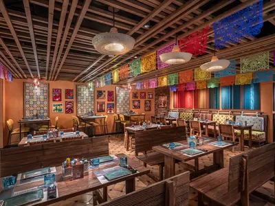 Der lebhafte Innenraum des mexikanischen Restaurants HOLA! Tacos & Cantina an Bord der MSC World Europa. Bunte Papel Picado-Banner und Sombrero-Lampen zieren die Holzdecke. Rustikale Holztische mit blauer Glasware stehen bereit für Gäste.