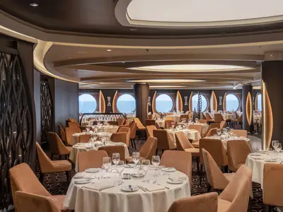 Elegantes Restaurant 'La Foglia' an Bord der MSC World Europa. Modern eingerichteter Raum mit braunen Stühlen, weißen Tischen und großen runden Bullaugen mit Meerblick. Leerer Raum mit gedämpftem Licht.