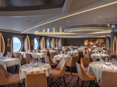 Elegantes Restaurant an Bord der MSC World Europa. Es zeigt zahlreiche eingedeckte Tische, braune Stühle und runde Fenster mit Meerblick. Die Einrichtung ist modern mit goldenen Blattmotiven.