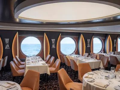 Ein elegantes Restaurant an Bord der MSC World Europa mit runden Bullaugen-Fenstern, die den Blick auf das blaue Meer freigeben. Die Tische sind gedeckt, orangefarbene Stühle stehen bereit.