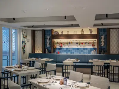 Ein helles, maritim eingerichtetes Restaurant, La Pescaderia, an Bord der MSC World Europa. Blaue Fliesen und nautische Dekorationen wie Muscheln, Bojen und ein Fischernetz praegen den Raum mit Tischen und Stuehlen.