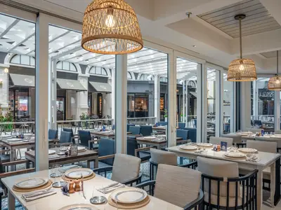 Blick in das helle und modern eingerichtete Spezialitätenrestaurant La Pescaderia an Bord der MSC World Europa. Mehrere gedeckte Tische und Stühle sind zu sehen, im Hintergrund weitere Geschäfte der Promenade durch Glasfenster.