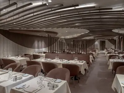 Der helle und weitläufige Speisesaal des Les Dunes Restaurants an Bord der MSC World Europa. Viele braune Sessel und Bänke stehen an weiss gedeckten Tischen. Die Decke ist mit geschwungenen Holzlamellen verziert, die Wände haben passende Holzpaneele.