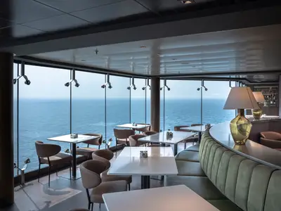 Elegantes Restaurant an Bord der MSC World Europa. Helle Tische und Stühle stehen vor bodentiefen Fenstern mit Meerblick. Eine geschwungene Bar mit Sitzbank und Tischlampen ist sichtbar. Ohne Personen.