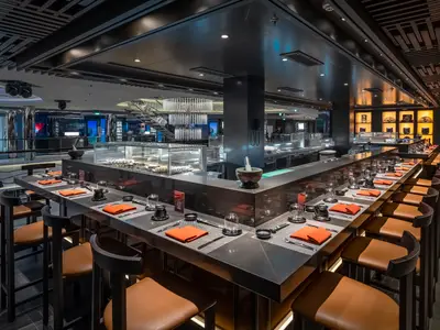 Einladende Kaito Sushi Bar an Bord der MSC World Europa. Die langgezogene Theke mit Hochstühlen ist elegant gedeckt, im Hintergrund ist die offene Küche sichtbar und ein Kronleuchter hängt über einer Treppe.