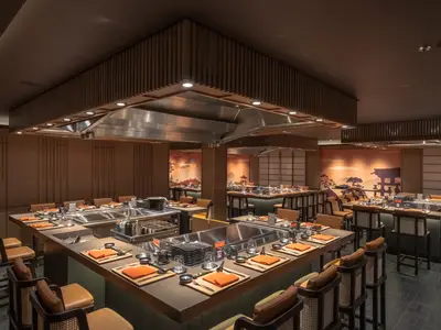 Das Kaito Teppanyaki Restaurant an Bord der MSC World Europa zeigt mehrere Kochstationen mit Sitzplätzen, japanischen Dekoren und warmer Beleuchtung. Die Tische sind mit orangefarbenen Servietten und Besteck gedeckt, bereit für Gäste. Keine Personen sind sichtbar.