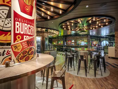 Blick in das lebhafte Pizza & Burger Restaurant an Bord der MSC World Europa. Einzigartige Wandgrafiken und neonbeleuchtete Essensmotive zieren den modernen Raum mit hohen Tischen und Barhockern.