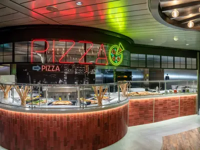 Ein modernes Pizza-Restaurant an Bord der MSC World Europa. Die rote Neonschrift 'PIZZA' dominiert über einer langen Buffettheke mit frischen Pizzen und Beilagen. Rote Ziegel und warmes Licht schaffen eine einladende Atmosphäre.