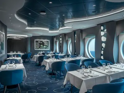An elegant specialty restaurant on board the MSC World Europa with blue chairs, set tables, and porthole windows offering ocean views. The ceiling features modern, wavy light installations, and the carpet has a circular pattern. No people are visible.