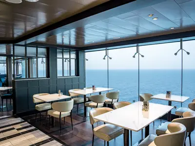 Blick in ein stilvolles Restaurant an Bord der MSC World Europa. Helle Tische und cremefarbene Stühle stehen vor einer großen Fensterfront, die eine weite Aussicht auf das blaue Meer bietet. Die Decke ist dunkel gehalten mit eingelassenen Lichtern.