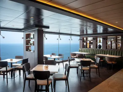 Elegantes Restaurant an Bord der MSC World Europa. Große Fenster bieten Panoramablick auf das Meer. Viele Tische und Stühle, grüne Sitzbänke und dekorative Regale prägen den Raum.