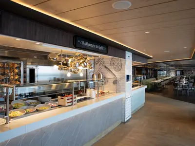 Ansicht des Il Mercato Buffets an Bord der MSC World Europa. Eine Rotisserie mit Hähnchen, eine große Salattheke und dahinter zahlreiche Esstische.