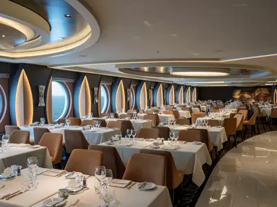 Elegantes Restaurant an Bord der MSC World Europa mit zahlreichen gedeckten Tischen und braunen Stühlen. Große Bullaugen mit Meerblick und moderne Lichtinstallationen prägen das stilvolle Ambiente.