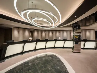Rezeption an Bord der MSC World Europa. Langer, geschwungener Tresen mit Beleuchtung und Monitoren. Futuristisches Deckenlicht, elegante Einrichtung, keine Personen.