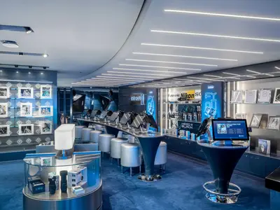 Moderner Foto-Shop 'The Hub' an Bord der MSC World Europa. Blaue Theke mit Bildschirmen und Hockern. Regale zeigen Kameras und Fotorahmen. Helle, moderne Beleuchtung.