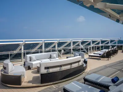 Sonnendeck der MSC World Europa mit modernen, hellen Loungemöbeln und Liegestühlen mit blauen Handtüchern. Glasbrüstung bietet weiten Meerblick unter blauem Himmel. Ohne Personen.