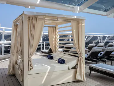Luxuriöse Cabana mit weissen Liegen und blauen Handtüchern auf dem sonnigen Deck der MSC World Europa. Im Hintergrund moderne Sonnenliegen und die Schiffsarchitektur unter klarem, blauem Himmel.
