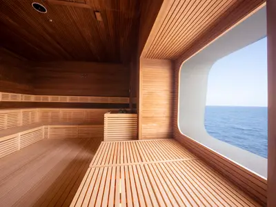 Eine helle, moderne Sauna an Bord der MSC World Europa. Die Wände und Sitzbänke bestehen aus hellem Holz. Rechts befindet sich ein großes, abgerundetes Fenster mit weitem Blick auf das blaue Meer und den Horizont unter einem klaren Himmel.