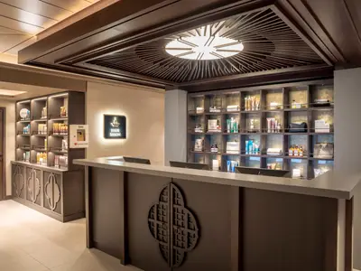 Empfangsbereich des eleganten Friseursalons an Bord der MSC World Europa. Ein dunkler Tresen mit Computermonitoren und ein Regal mit Haarprodukten sind sichtbar. Ein Defibrillator ist an der Wand montiert.