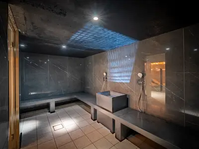 Modernes Dampfbad im MSC Aurea Spa der MSC World Europa. Es zeigt dunkle Marmorwände, Betonbänke, ein Wasserbecken, Duschbrausen und eine Gitter-Lichtprojektion an der Decke.