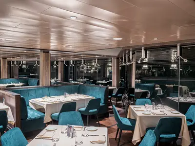 Ein elegantes Restaurant an Bord der MSC World Europa. Der Raum ist mit teal-blauen Polsterbänken und stilvoll gedeckten Tischen ausgestattet. Moderne Beleuchtung und Spiegel tragen zur luxuriösen Atmosphäre bei.
