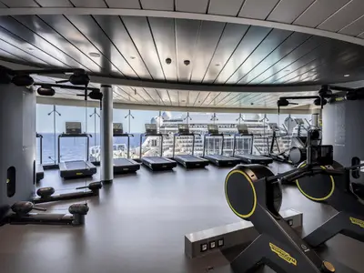 Ein modernes Fitnessstudio an Bord der MSC World Europa mit einer Reihe von Technogym-Laufbändern und Rudergeräten vor großen Fenstern mit Meerblick.