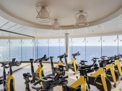 Moderner Fitnessraum der MSC World Europa mit gelben Technogym-Spin-Bikes. Raumhohe Fenster bieten weiten Meerblick. Helle Decke mit Design-Pendelleuchten und Holzfußboden. Offen und sportlich.