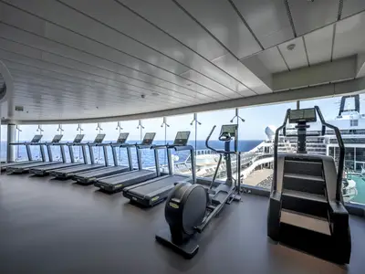 Modernes Fitnessstudio auf der MSC World Europa mit Laufbändern, Ellipsentrainer und Stairclimber. Große Fenster bieten Meerblick und zeigen die Außendecks des Schiffes.