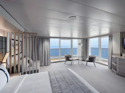 Eine helle, modern eingerichtete Grand Suite Aurea mit Balkon an Bord der MSC World Europa. Ein Doppelbett, eine Sitzecke und ein Meerblick durch die bodentiefen Fenster sind zu sehen.