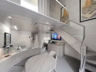 Eine moderne, hell gestaltete Duplex Suite an Bord der MSC World Europa. Im Vordergrund ein runder Esstisch mit Sitzbank. Dahinter ein Wohnbereich mit Sofa, TV, Minibar und einer Treppe zur oberen Schlafebene.