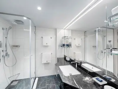 Modernes Badezimmer an Bord der MSC World Europa mit zwei Waschbecken, einer geräumigen Glasdusche und eleganten Marmorflächen. Weiße Handtücher mit Logo sind bereitgelegt.