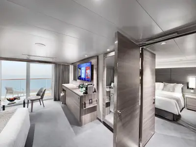 Eine helle, moderne Suite mit Balkon an Bord der MSC World Europa. Man sieht den Wohnbereich mit Sofa, Tisch und Fernseher sowie den Zugang zum Schlafzimmer. Draußen ist das Meer sichtbar.