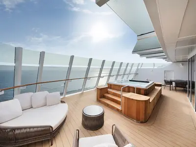 Die große, private Terrasse einer Owner's Suite auf der MSC World Europa bietet einen Whirlpool, eine Loungeliege und Esstische mit Meerblick unter klarem Himmel.