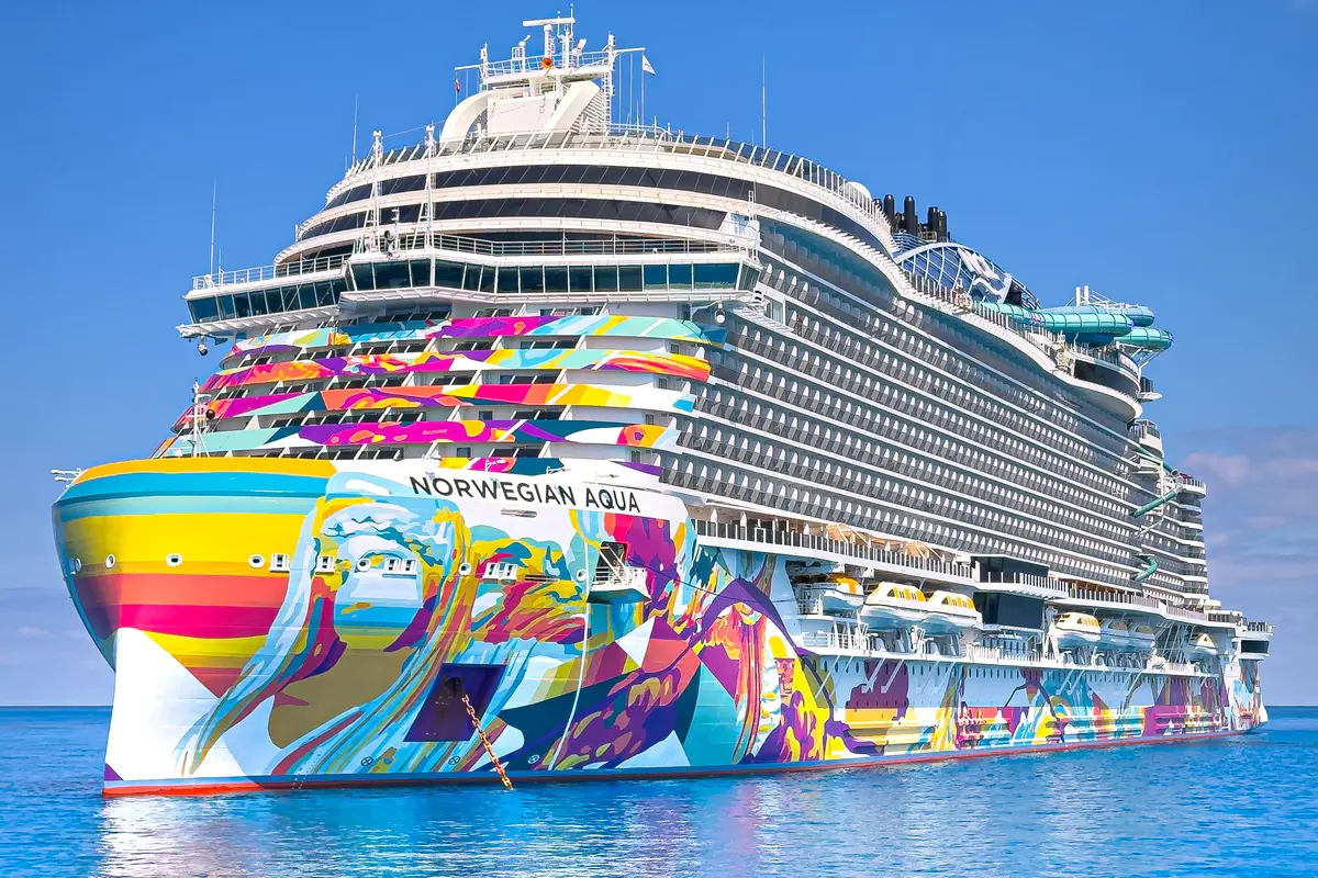 Die Norwegian Aqua, ein großes Kreuzfahrtschiff, fährt auf blauem Wasser. Das Schiff hat eine auffällige, farbenfrohe Kunst am Rumpf mit abstrakten Mustern und Gesichtern. Im Hintergrund ist ein klarer blauer Himmel zu sehen.