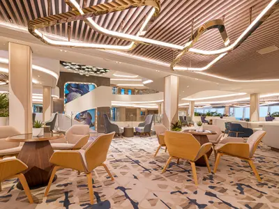 Modernes Atrium an Bord der Norwegian Aqua mit stilvollen Sitzgruppen. Auffällige Kunst auf einer geschwungenen Treppe und Meerblick.