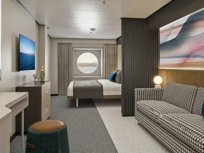 Eine elegant eingerichtete Aussenkabine an Bord der Norwegian Aqua. Im Vordergrund ein Sofa mit Hahnentrittmuster, dahinter ein Doppelbett und ein rundes Fenster mit Meerblick.