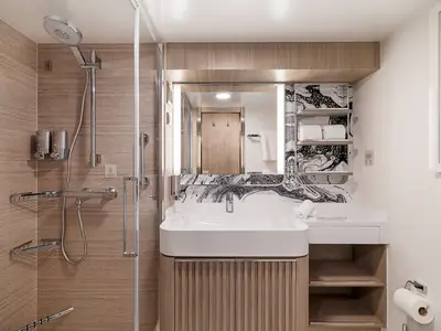 Detailansicht eines modernen Badezimmers an Bord der Norwegian Aqua. Links die Duschkabine, daneben ein Waschbecken mit beleuchtetem Spiegel und Ablagen.