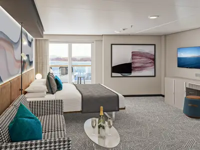 Eine elegante Club Balcony Suite an Bord der Norwegian Aqua. Sie zeigt ein Doppelbett, eine Couch, einen Couchtisch mit Sekt, einen Schminktisch und einen Balkon mit Meerblick.